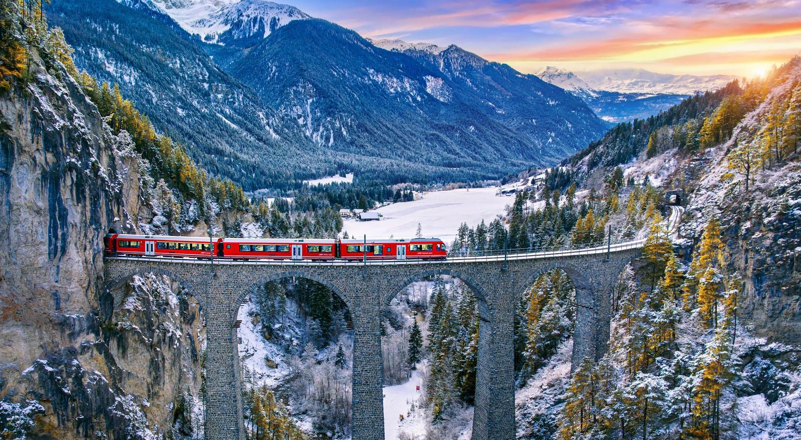 Red train traveling across the Landwasser Viaduct in the Swiss Alps surrounded by green forests and mountains under a clear sky, conveying a sense of adventure and tranquility
